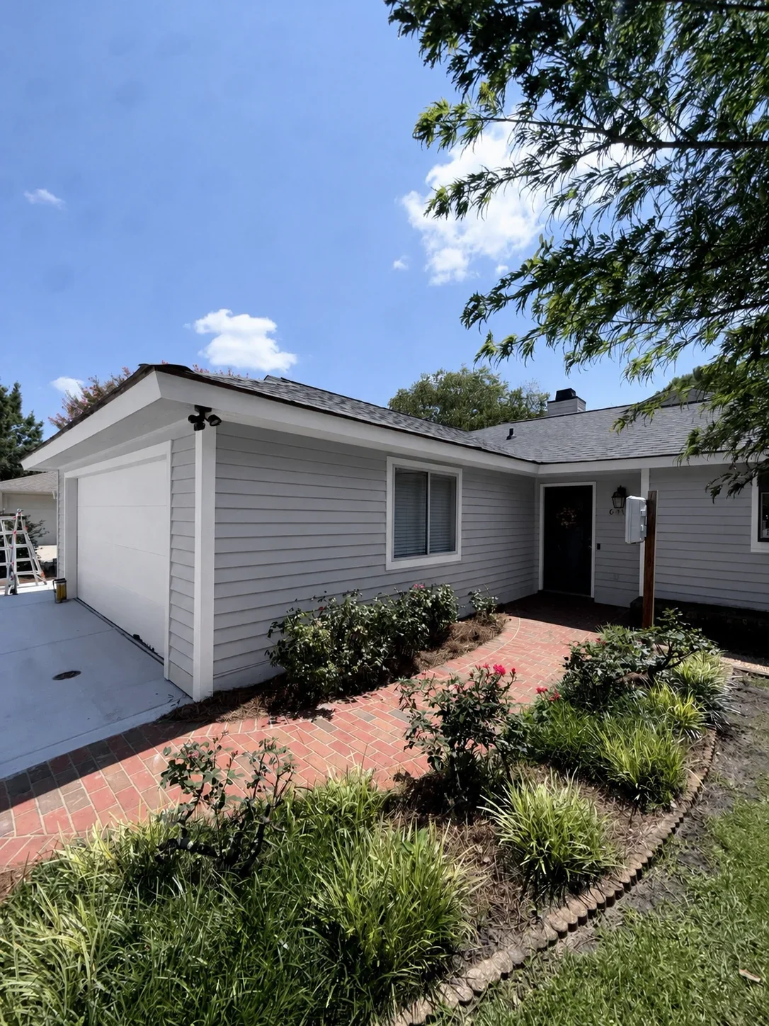 Light gray horizontal siding with white trim, black roof, brick paver walkway, and landscaped garden beds