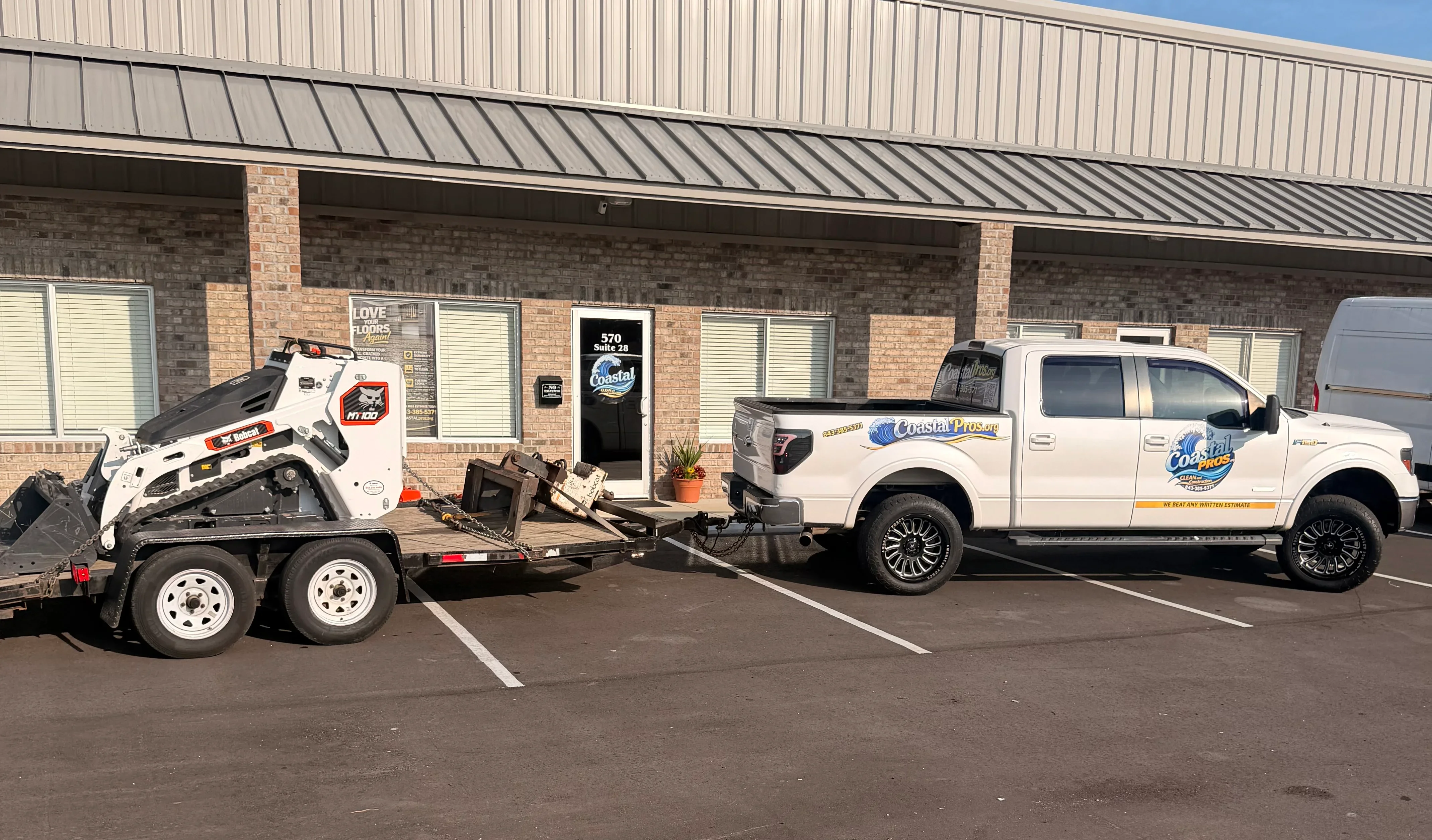 White dual-axle trailer with skid steer loader and service truck parked at commercial facility